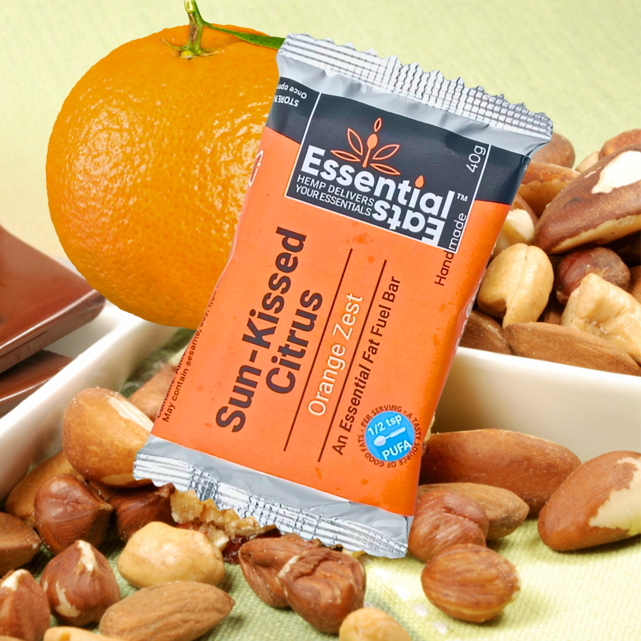 Essentialerna snack bar with sun-kissed citrus flavor surrounded by nuts and an orange on a green surface.